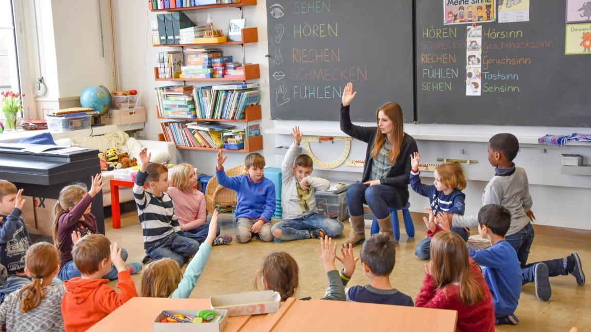 Eine Unterstufenschulklasse im Sitzkreis vor der Wandtafel am Abstimmen