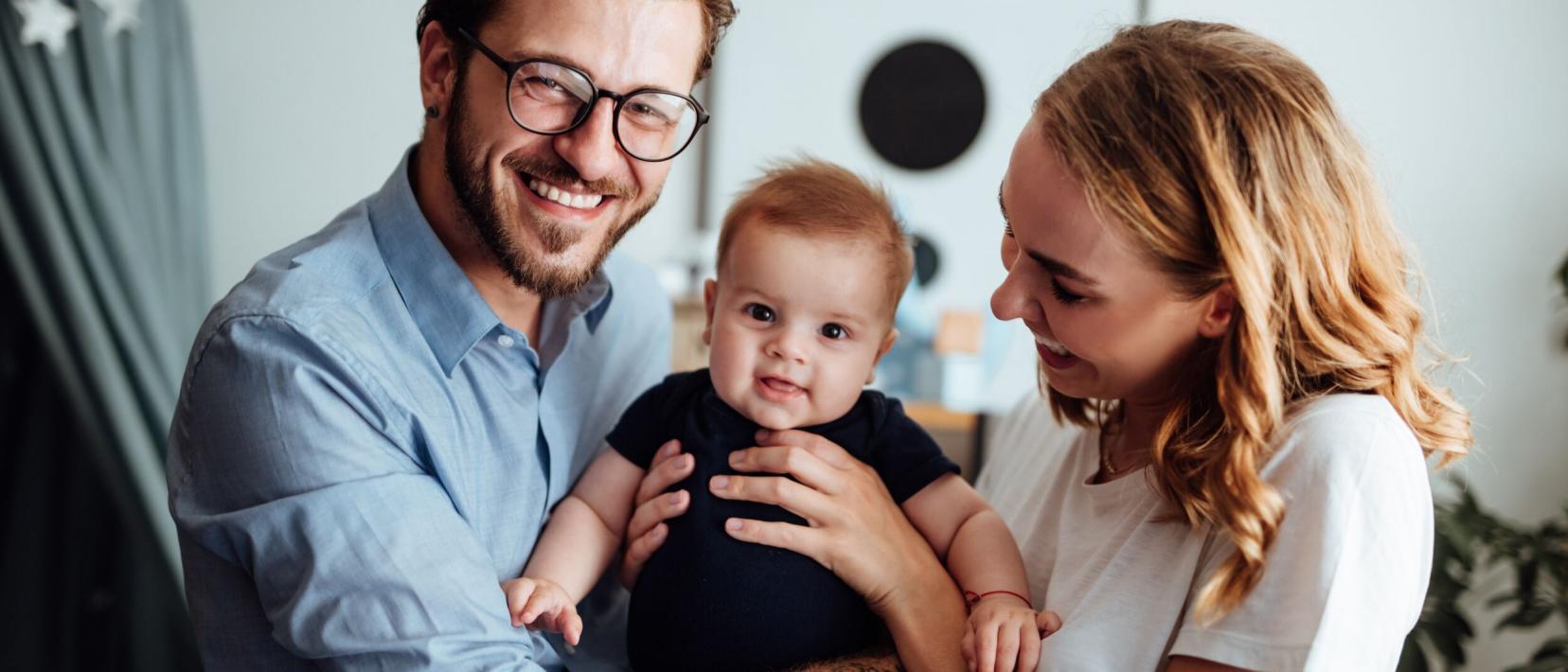 Ein Mann und eine Frau halten strahlend ein lächelndes Baby in die Kamera.