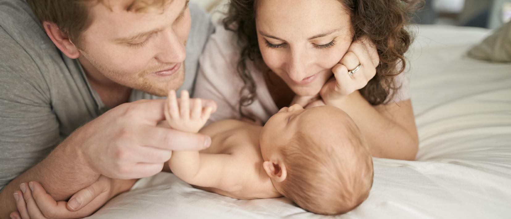 Eine Frau und eine Mann liegen bäuchlings auf einem weissen Laken und schauen liebevoll auf ein Baby, dass neben ihnen liegt.