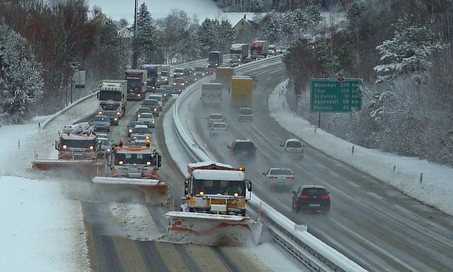 Drei grosse Fahrzeuge mit Pfadschlitten räumen gestaffelt eine Autobahn; dahinter gibt es dadurch einen langen Rückstau.