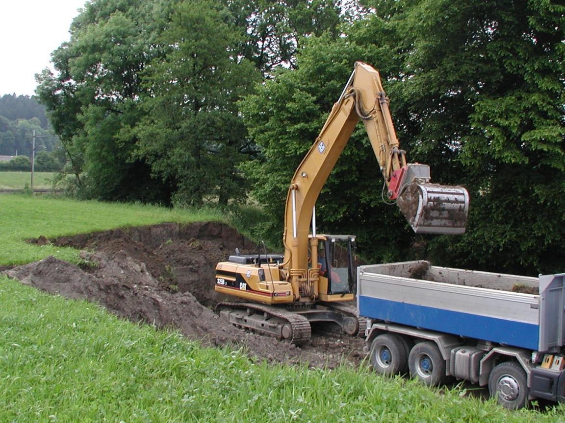 Ein Bagger trägt an einer Wiese Boden ab und belädt damit einen Lastwagen.