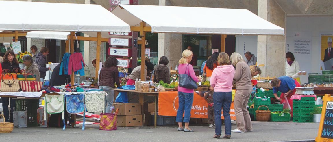 Wochenmarkt im Ortszentrum von Meilen. Das Bild wird geprägt von zwei Markständen mit weissem Dach. Auf dem linken werden Textilwaren, auf dem rechten exotische Früchte angeboten. Hinter den Tischen Marktfahrerinnen, davor eine Gruppe von drei Frauen und einem Kind.