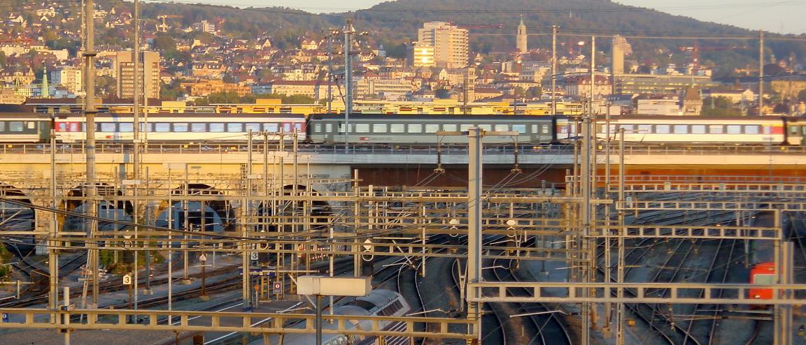 Gleisfeld im goldenen Abendlicht bei der Einfahrt zum Hauptbahnhof Zürich. Im Vordergrund ein Gewimmel von unzähligen Strommasten, in der Bildmitte ein Zug, der auf dem Lettenviadukt das Gleisfeld quert, im Hintergrund der Zürichberg.