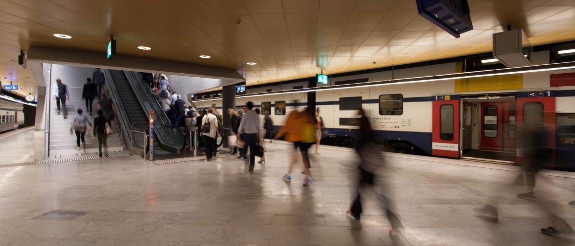 Morgendliche Pendler hetzen durch den unterirdischen Bahnhof Löwenstrasse im Zürcher Hauptbahnhof. Auf beiden Seiten stehen Züge, in der Bildmitte streben verschwommene Gestalten auf Treppe und Rolltreppe zu.