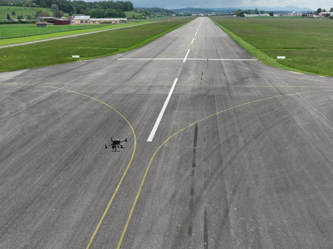 Quadkopter überfliegt eine asphaltierte Piste. Links und rechts davon befindet sich kurzgeschnittene Wiese, wenige Bäume und ein paar Gebäud ein der Ferne.